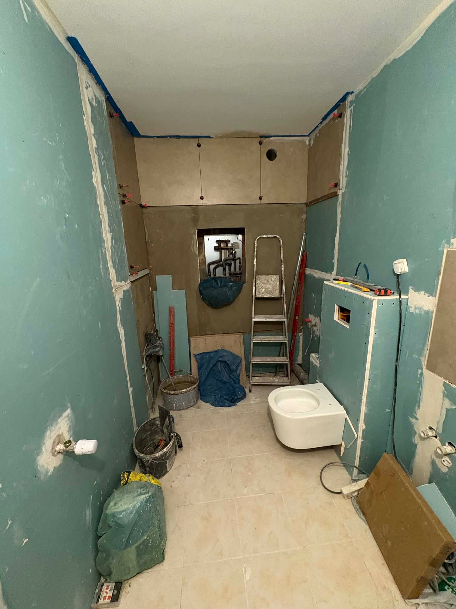 Łazienka w trakcie remontu: ściany z płyt karton-gips, częściowo położone płytki, zamontowana toaleta podwieszana, drabina i narzędzia. Widoczne ślady szpachlowania.