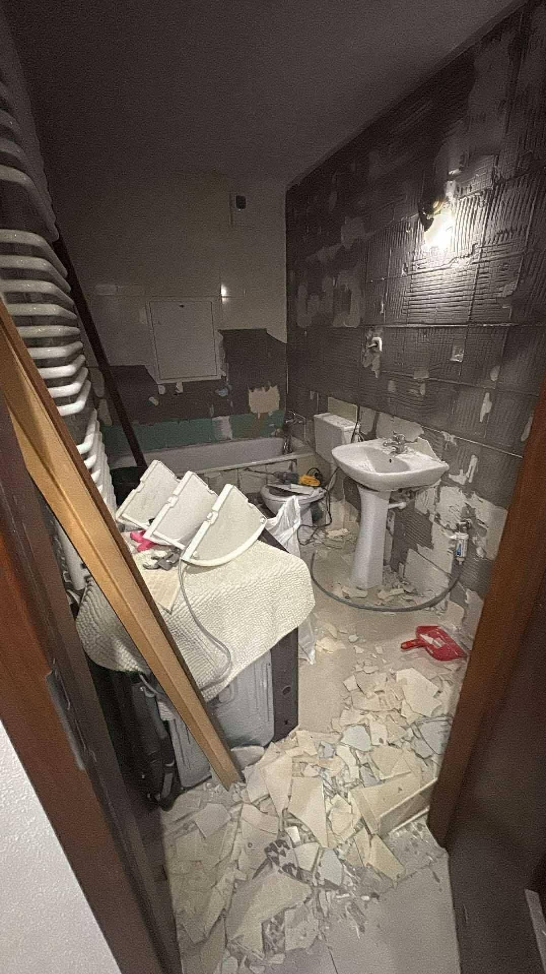 Łazienka w trakcie demontażu płytek, widoczne gruzowisko, umywalka na postumencie, wanna i narzędzia. Przygotowanie do remontu, ciemne płytki częściowo zerwane.