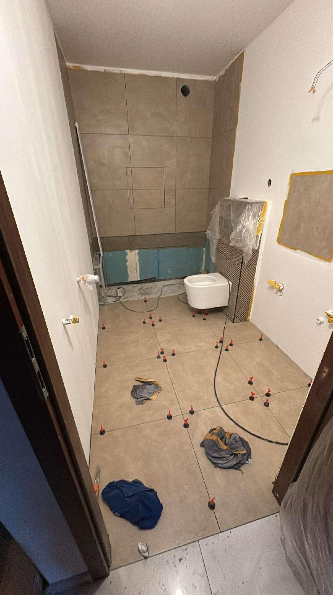 Łazienka w trakcie układania płytek podłogowych, widoczne poziomice i system poziomowania płytek. Częściowo wykończone ściany z zamontowaną toaletą pod folią.