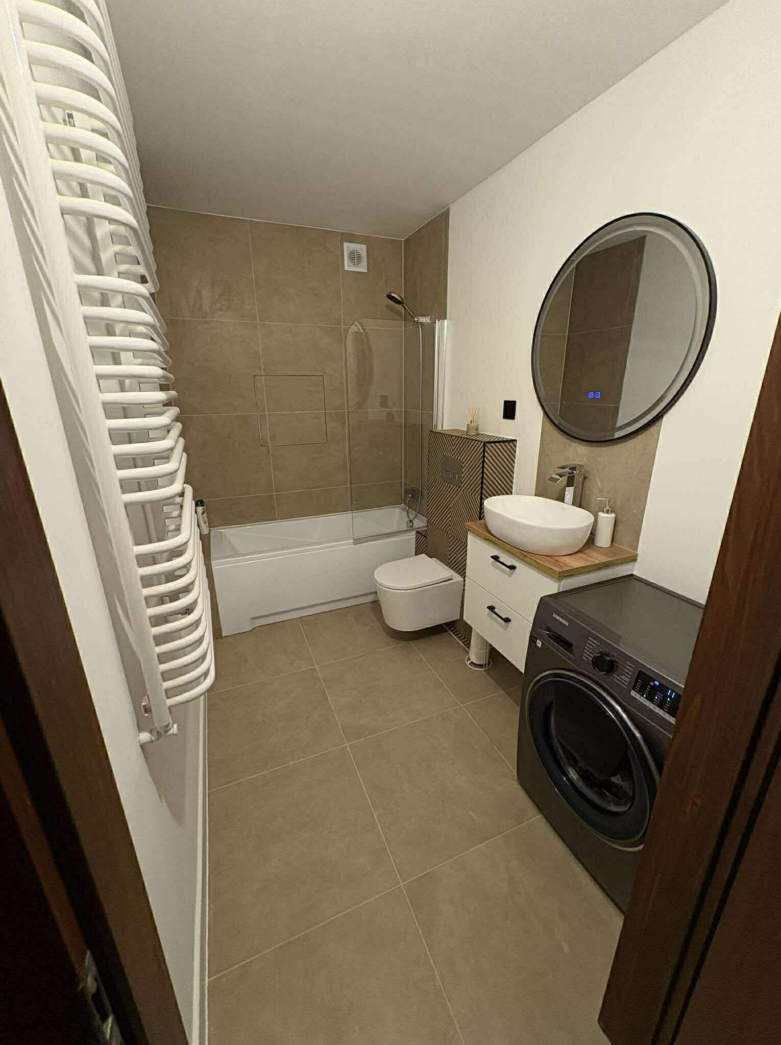 Nowoczesna łazienka w beżach: wanna z parawanem, umywalka na szafce z drewnianym blatem, okrągłe lustro i pralka. Grzejnik drabinkowy po lewej.