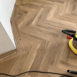 SZULC GROUP MIKOŁAJ SZULC - Ułożone panele podłogowe w jodełkę, widoczna listwa przypodłogowa i odkurzacz. Fragment podłogi z drobnymi zabrudzeniami po montażu.