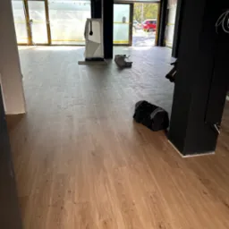 Jasne panele podłogowe w komercyjnym wnętrzu, widok z góry. Przy czarnej kolumnie leży czarna torba sportowa. Przez okno widać zieleń.