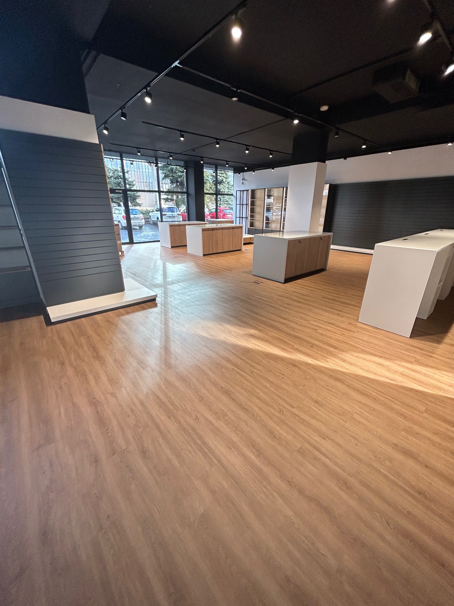 Jasne panele podłogowe w komercyjnej przestrzeni z oknami, oświetleniem punktowym i minimalistycznymi meblami. Widok na zewnątrz przez okna.