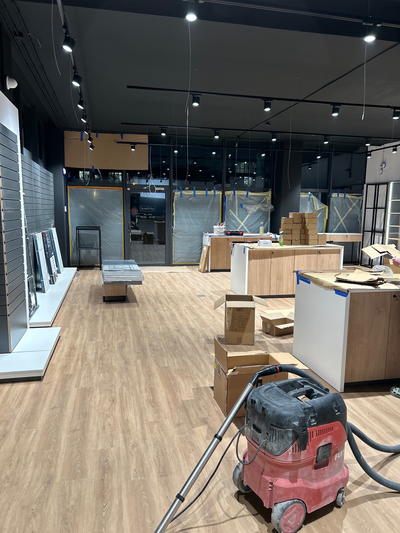 Montaż mebli sklepowych w Łodzi, widok na posadzkę i odkurzacz przemysłowy. Sklep w trakcie wykańczania, witryny zabezpieczone folią, kartony i narzędzia.