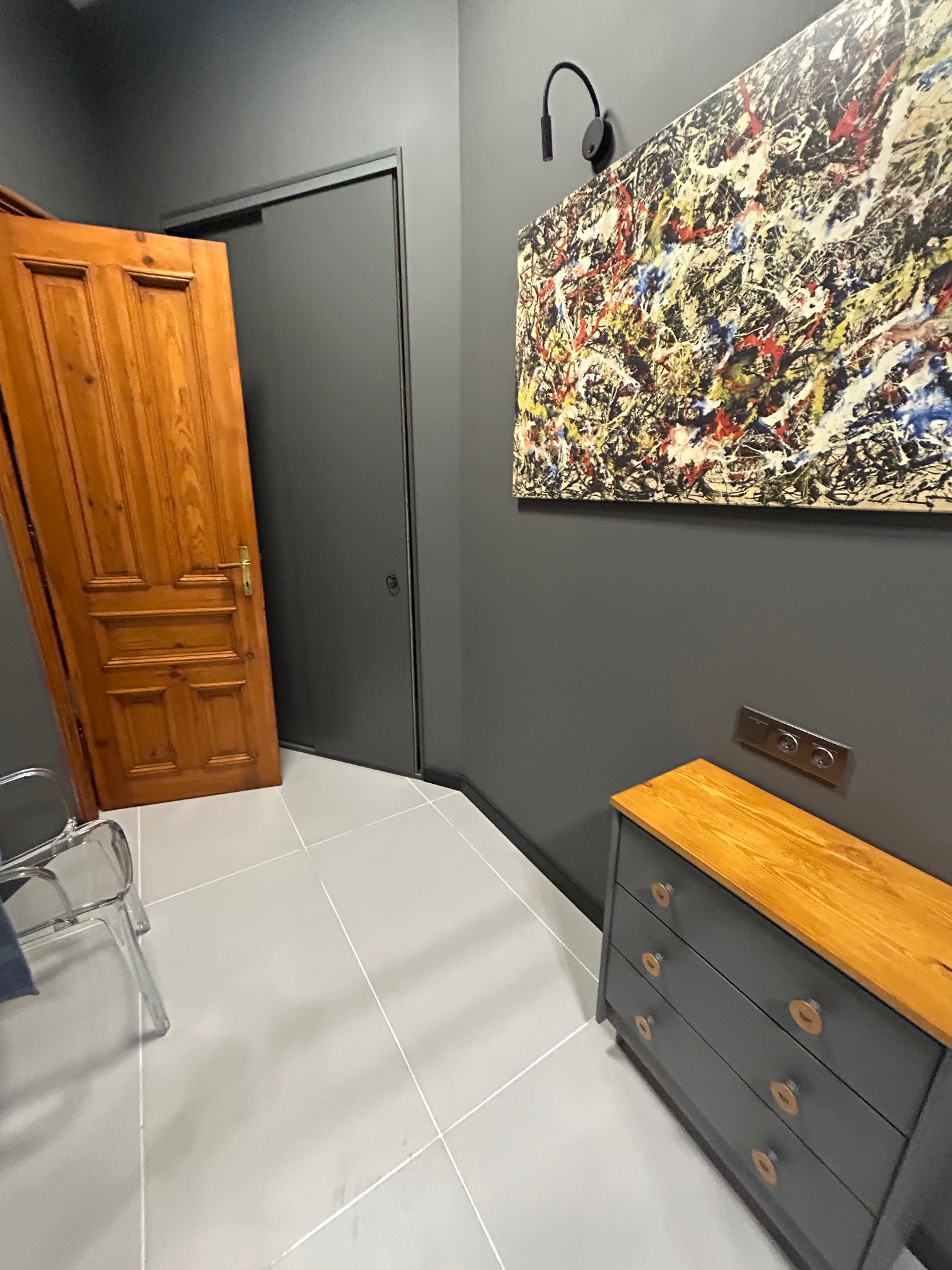 Szare wnętrze z drewnianą komodą i obrazem w stylu Pollocka. Widoczne drzwi drewniane i przesuwne, szare ściany oraz podłoga z jasnych płytek.