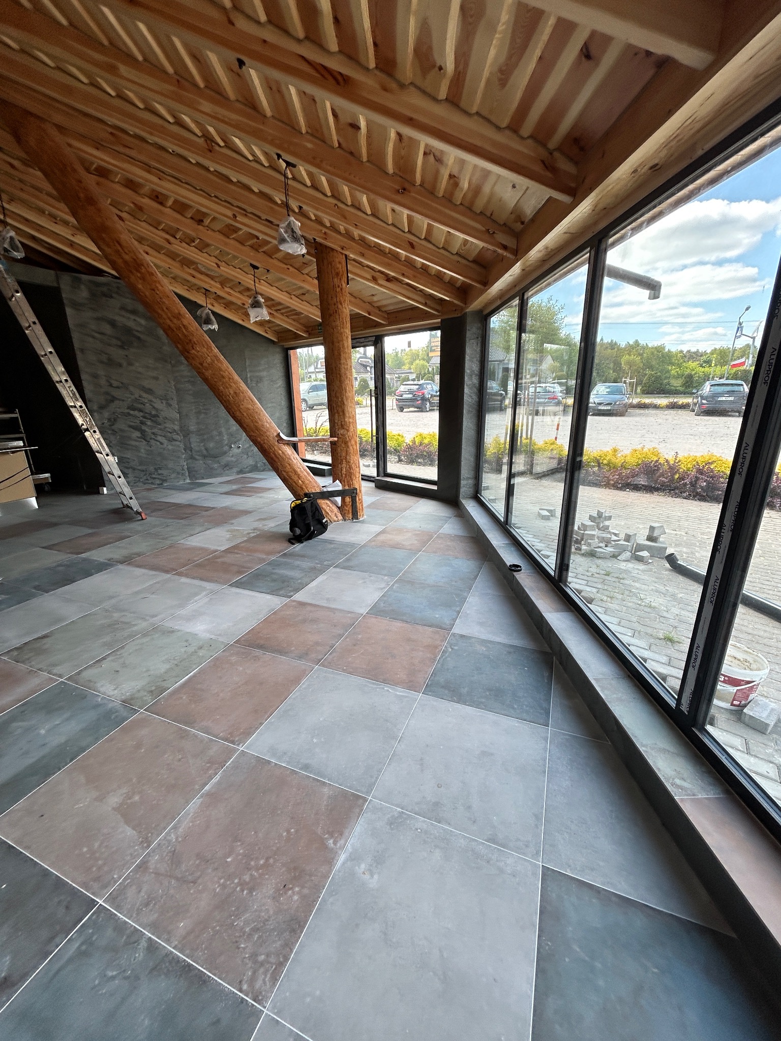 Wnętrze z drewnianą konstrukcją dachu i dużymi oknami. Na podłodze ułożona jest kolorowa, kwadratowa terakota. Widoczna drabina i torba narzędziowa.