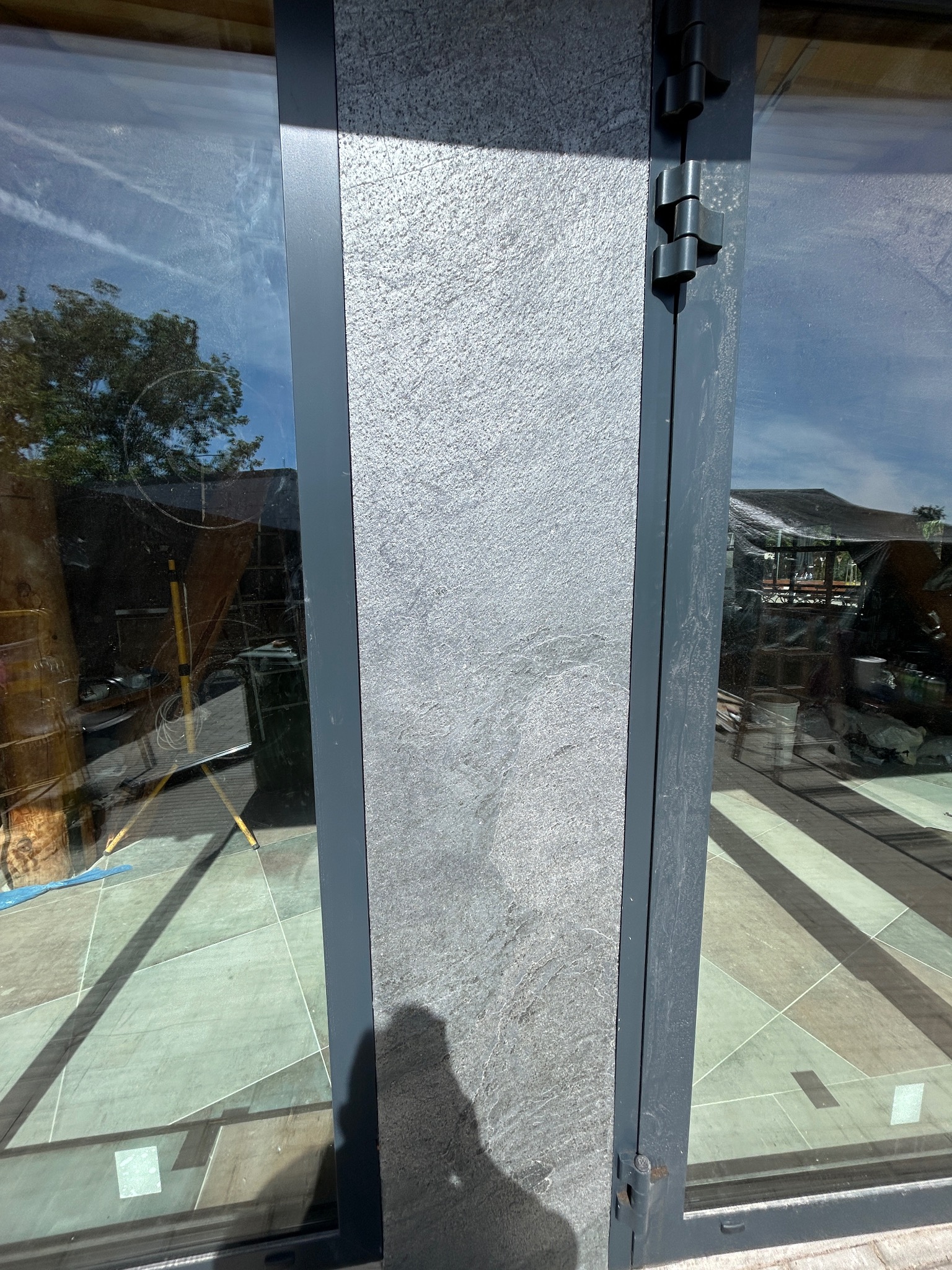 Szczegółowe ujęcie fragmentu ściany z szarego kamienia dekoracyjnego, przylegającego do okna z szarą ramą i widocznymi zawiasami. W tle odbicie otoczenia.