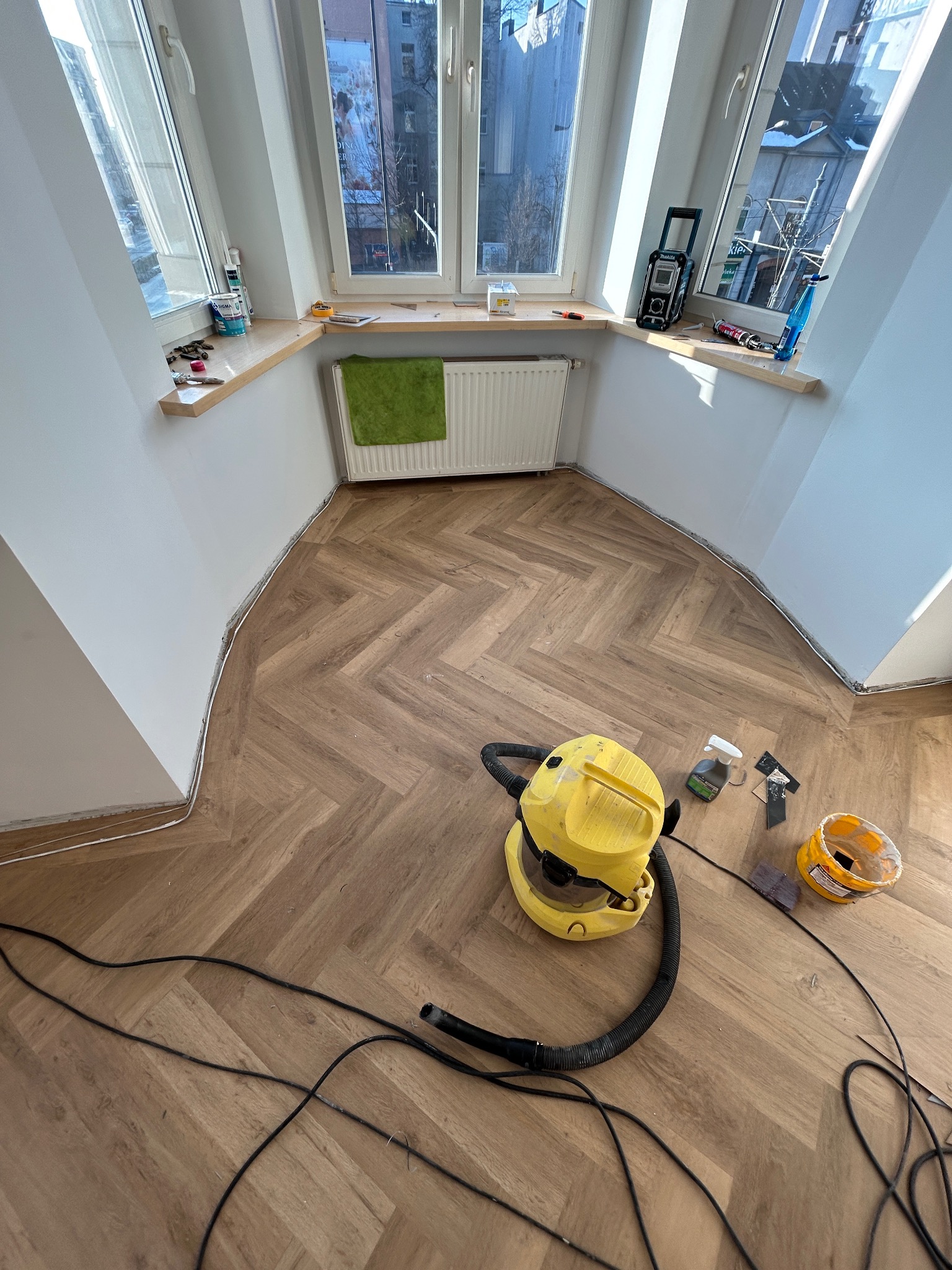 Ukończone układanie parkietu w jodełkę w pokoju z oknami, widoczny odkurzacz i akcesoria do sprzątania, białe ściany i parapety, widok na budynki z zewnątrz.