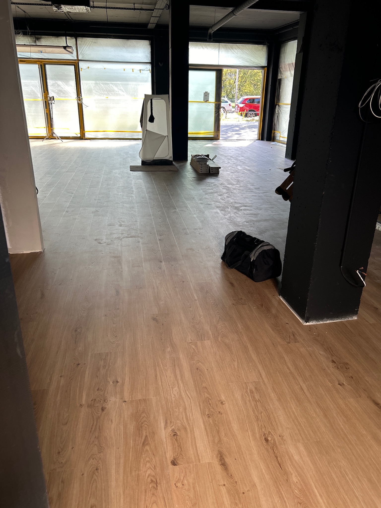 Jasne panele podłogowe w komercyjnym wnętrzu, widok z góry. Przy czarnej kolumnie leży czarna torba sportowa. Przez okno widać zieleń.