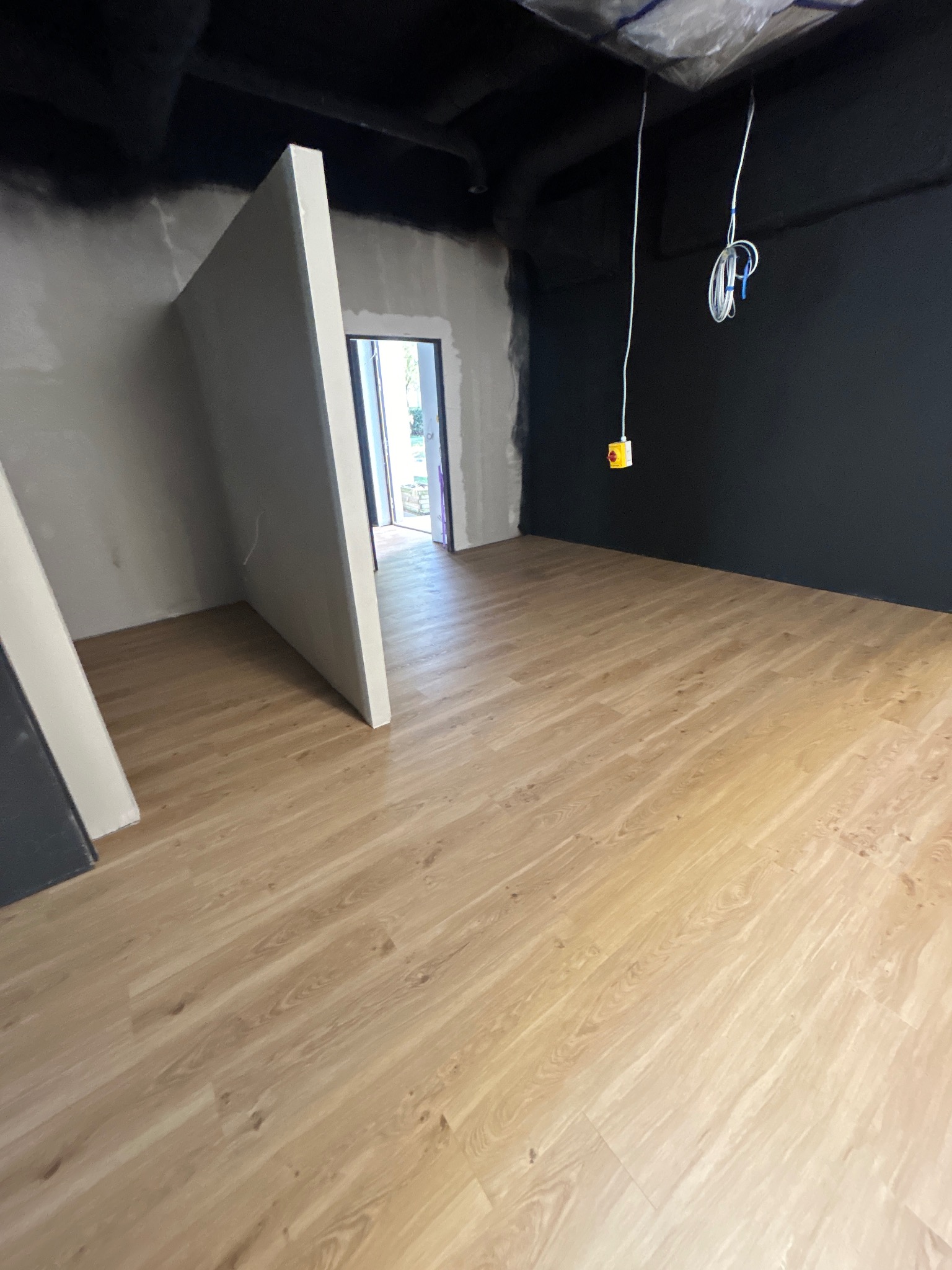 Jasne panele podłogowe w nowym wnętrzu, ściany w szarości i czerni, widoczne przewody elektryczne. Wykończenie podłogi w trakcie remontu.