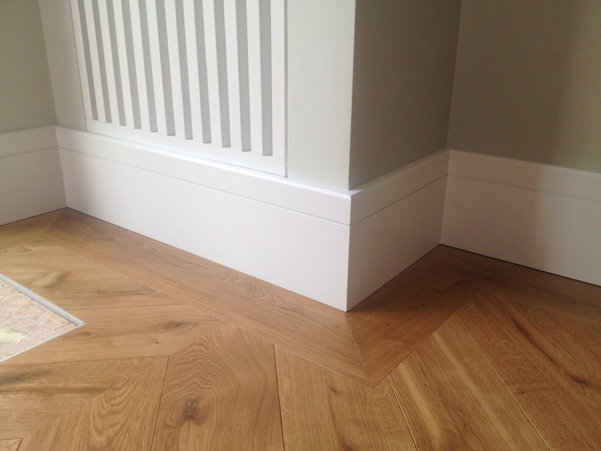 Parkiet ułożony we wzór jodełki, białe listwy przypodłogowe przy ścianie i grzejniku. Detal wykończenia wnętrza.