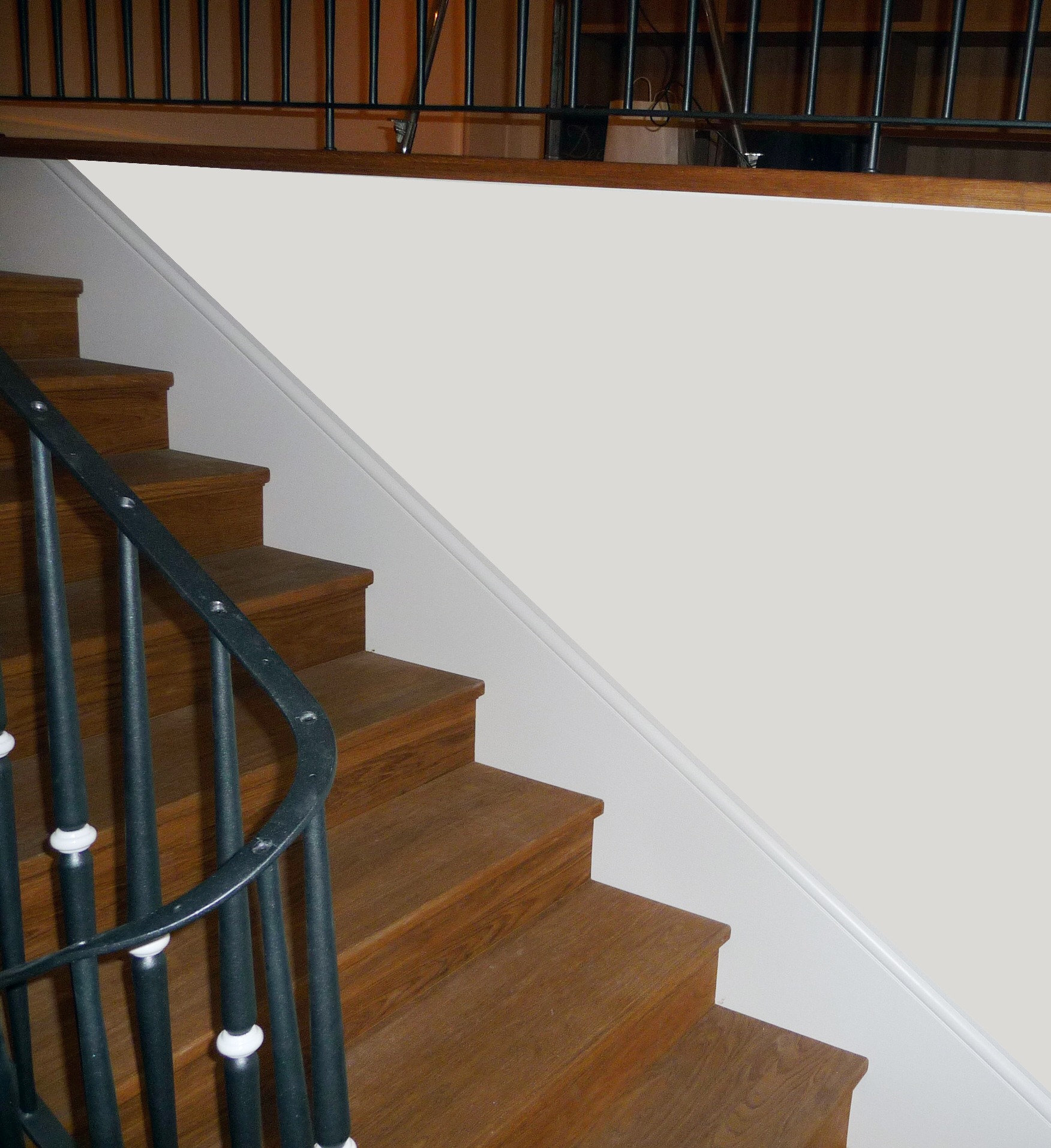 Drewniane schody z czarną, metalową balustradą i białymi akcentami, prowadzące na wyższą kondygnację. Ściana pomalowana na biało.