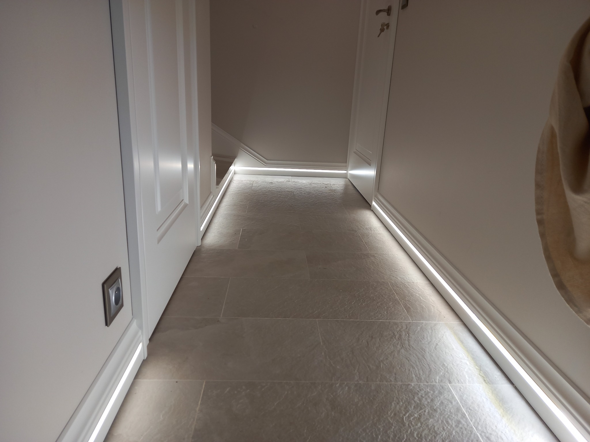 Korytarz z szarą kamienną podłogą i listwami przypodłogowymi LED, oświetlającymi przejście. Widoczny włącznik na ścianie i fragment wieszaka na ubrania.