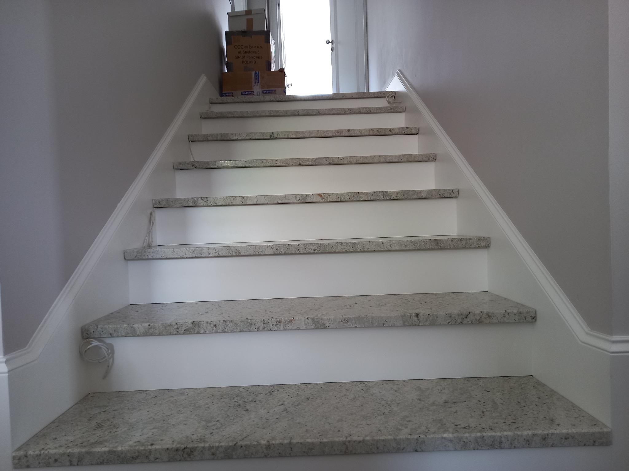 Wykończone schody wewnętrzne z szarymi stopniami i białymi podstopnicami. Na górze schodów leżą kartony. Widok z dołu.