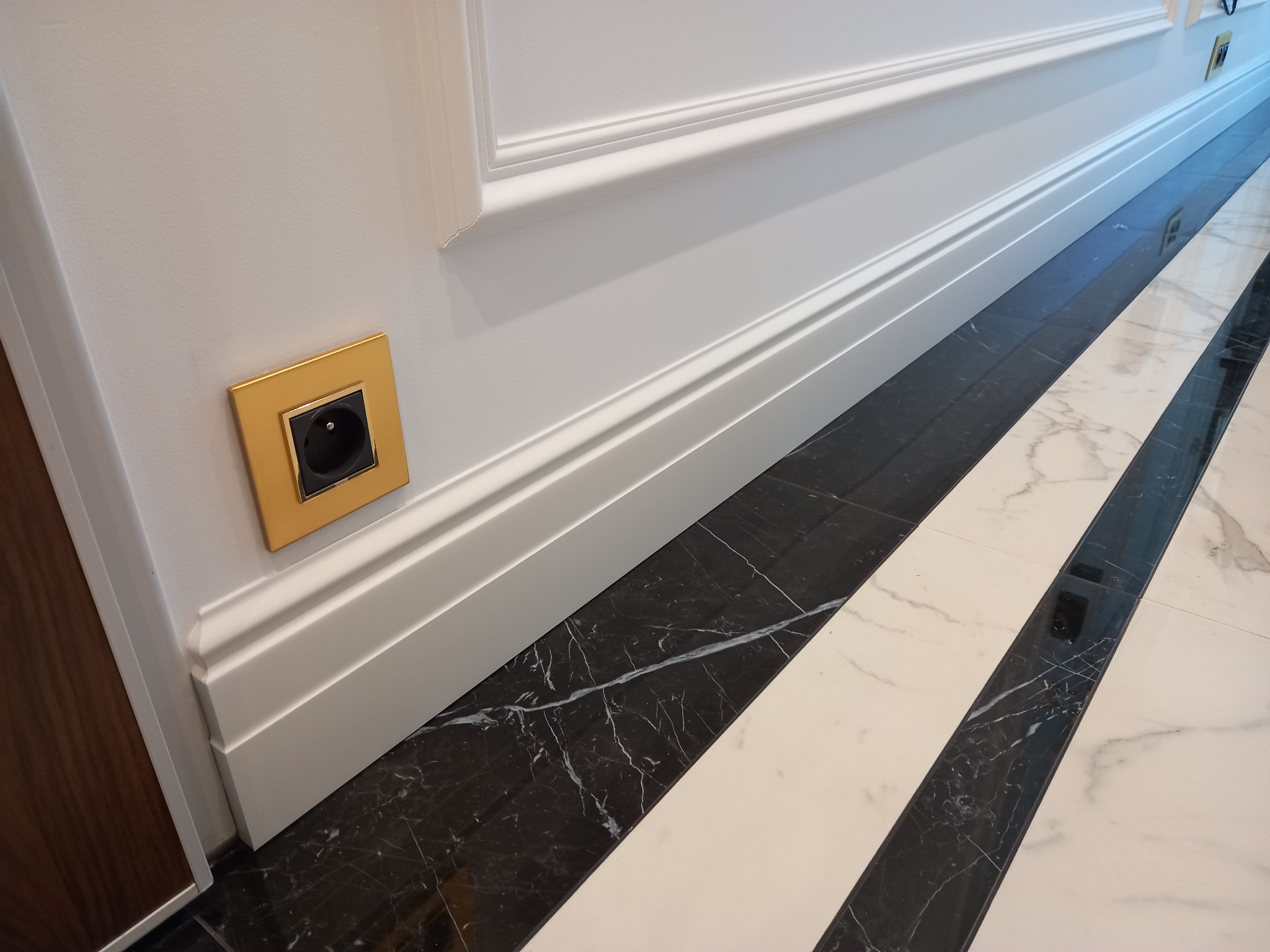 Biała ściana z dekoracyjną listwą i złotym gniazdkiem elektrycznym. Na podłodze wzór z czarno-białych marmurowych pasów. Detal wykończenia wnętrza.