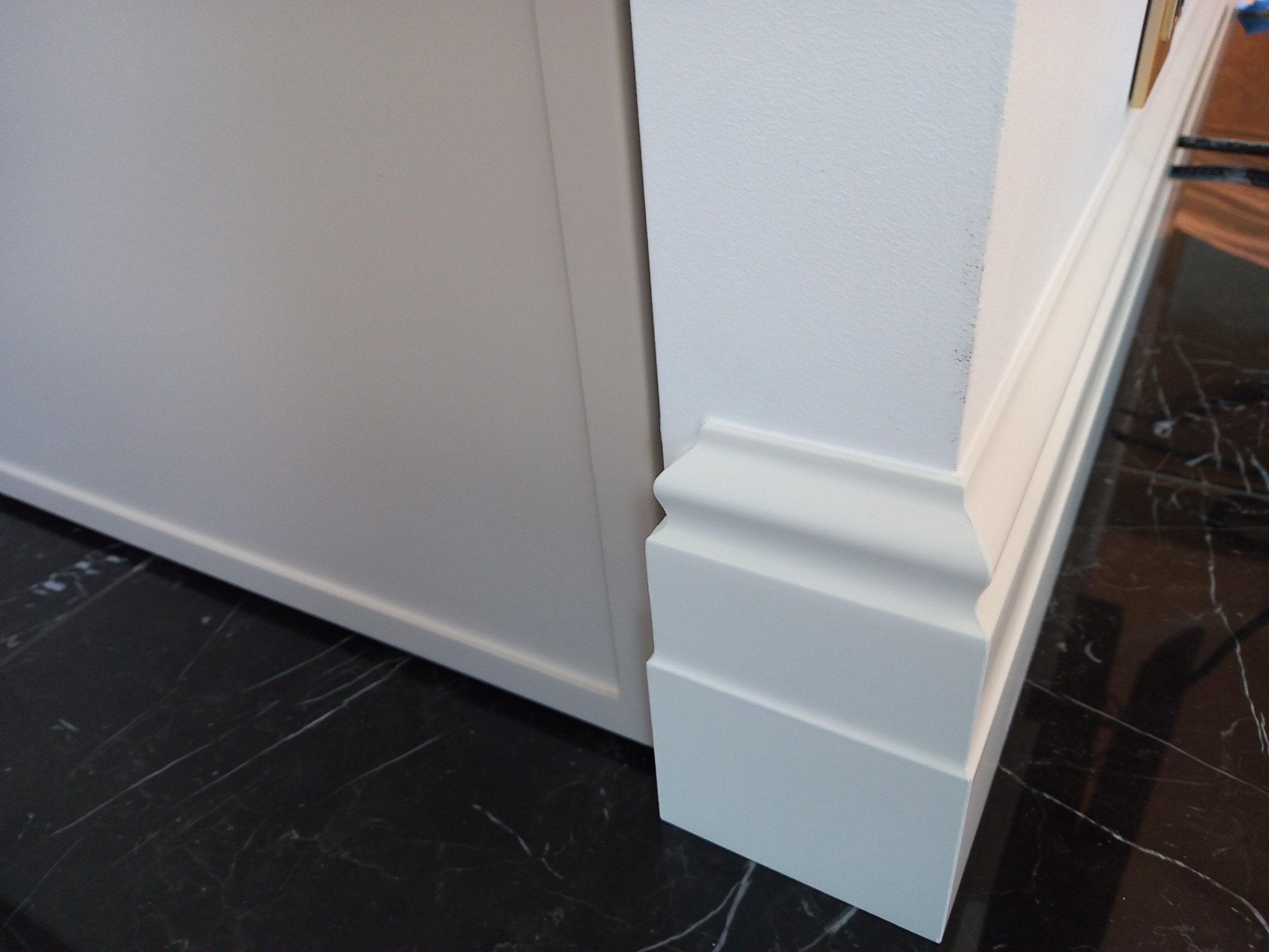 Biała listwa przypodłogowa z dekoracyjnym frezem, kontrastująca z ciemną, marmurkową podłogą. Element wykończenia wnętrza, widoczny fragment ściany i szafki.