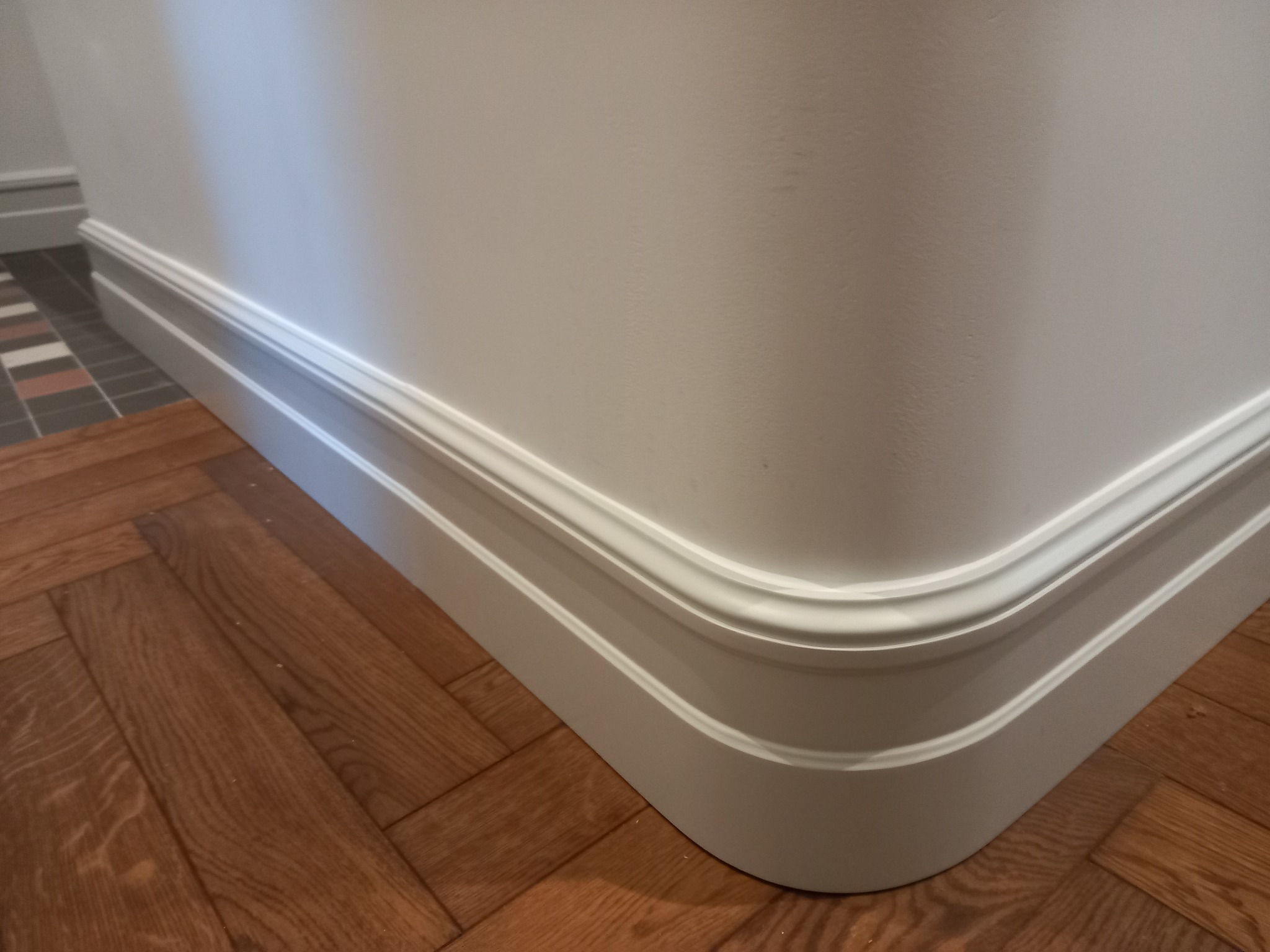 Biały narożnik ściany z listwą przypodłogową i parkietem ułożonym w jodełkę francuską. Fragment podłogi z płytkami w tle.