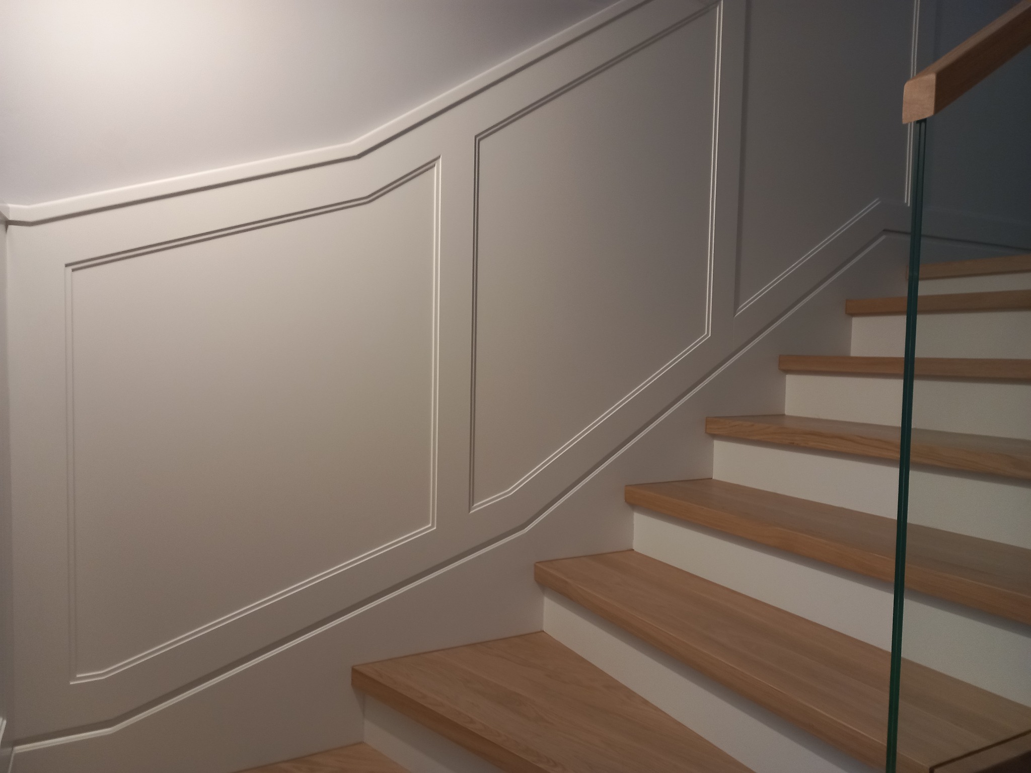 Eleganckie wykończenie ściany schodów z panelami dekoracyjnymi i drewnianymi stopniami. Minimalistyczny design z elementami szkła.