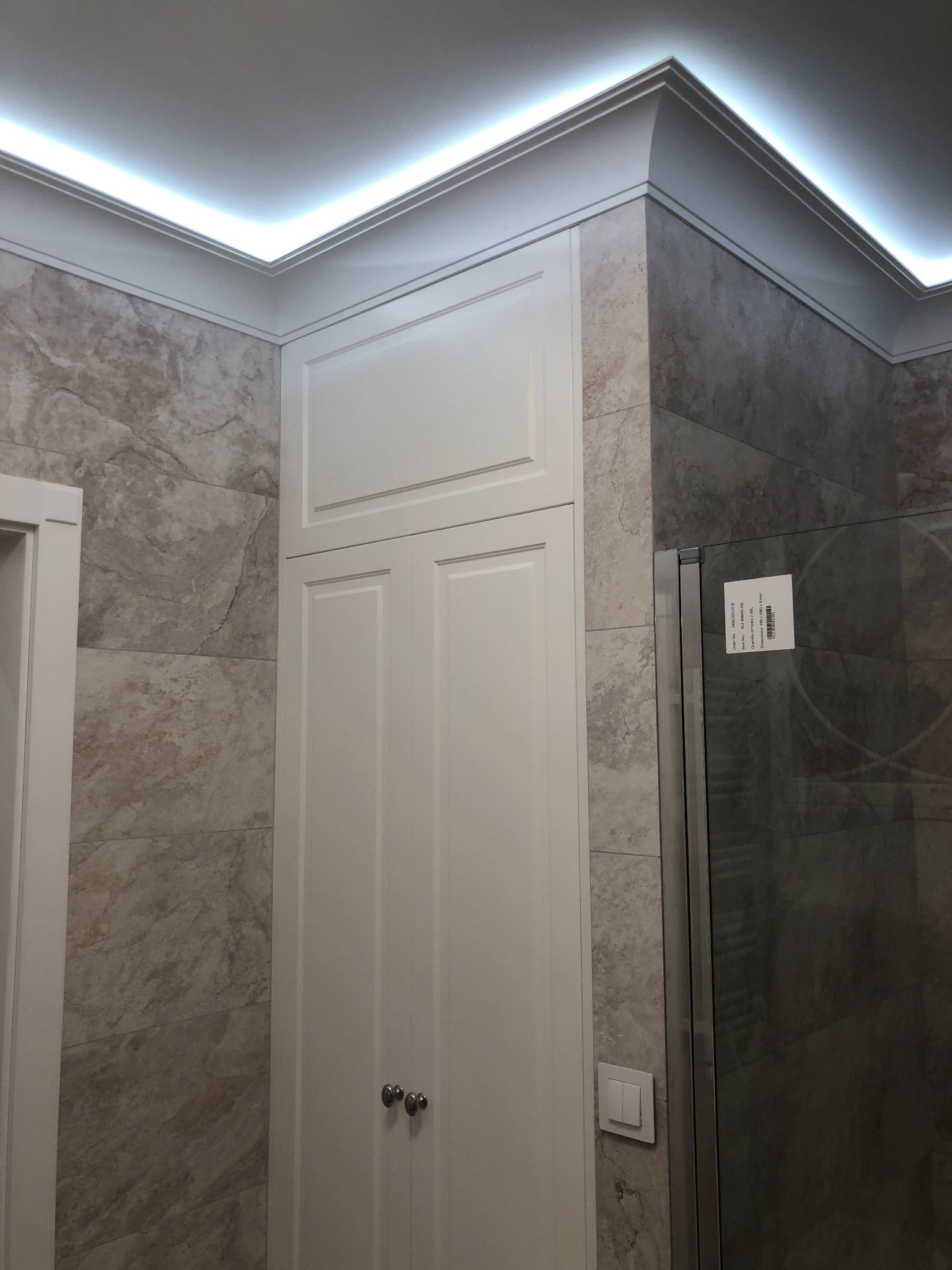 Elegancka zabudowa w łazience z białymi szafkami, marmurowymi płytkami i subtelnym oświetleniem LED na suficie, tworząca spójną i estetyczną całość.