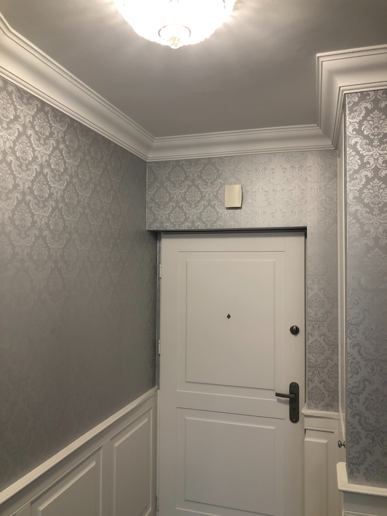 Eleganckie wykończenie przedpokoju: wzorzysta tapeta w odcieniach szarości, białe listwy przypodłogowe i sufitowe. Białe drzwi wejściowe z ciemną klamką.