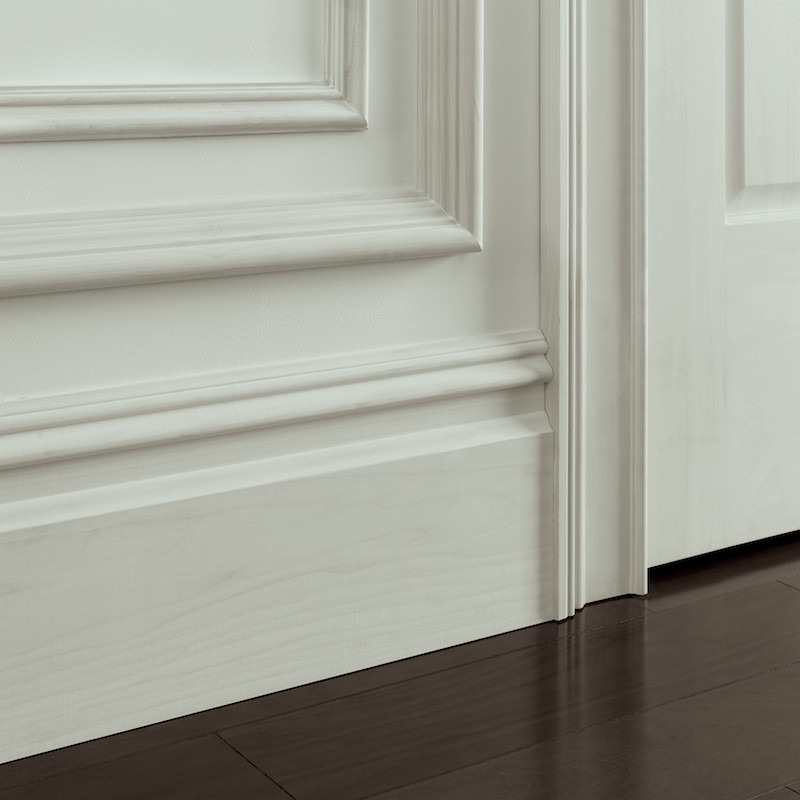 Biała, profilowana listwa przypodłogowa i panel ścienny z dekoracyjnymi frezami, przylegające do ciemnej drewnianej podłogi i białych drzwi.
