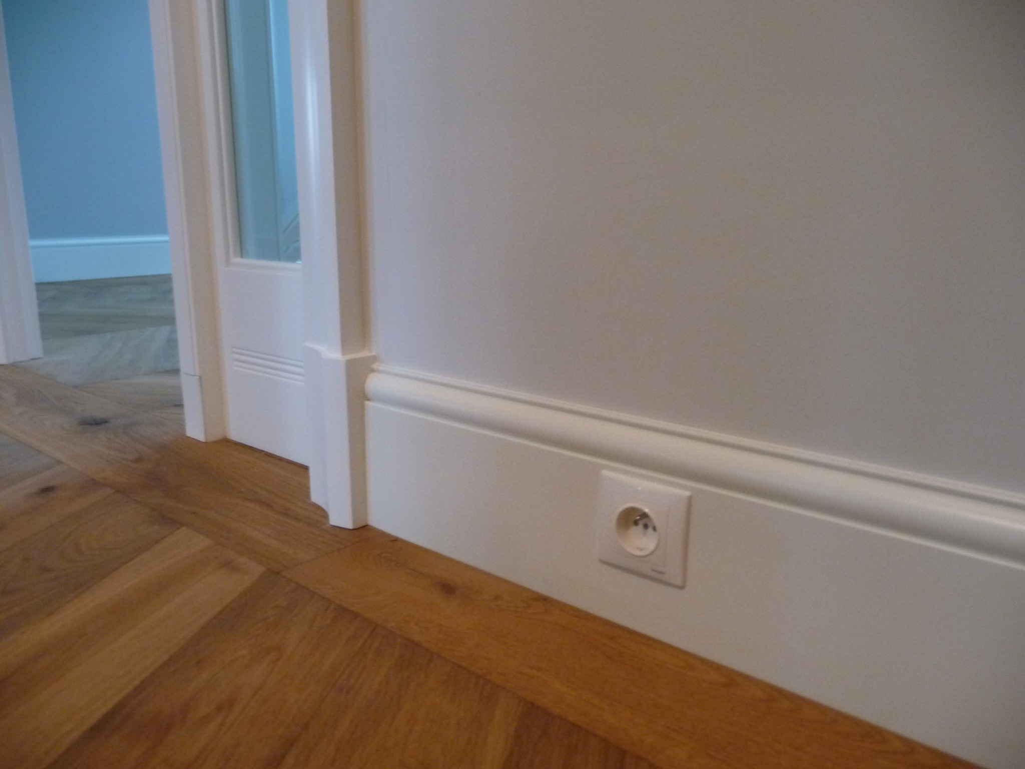 Biała ściana z listwą przypodłogową i gniazdkiem elektrycznym, parkiet ułożony w jodełkę, fragment białych drzwi. Detal wykończenia wnętrza w jasnej tonacji.