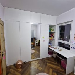 Błażej Michalak - Biała szafa wnękowa z lustrem i biurkiem w pokoju dziecięcym. Podłoga w jodełkę, dekoracje na ścianach. Widok na wnętrze pokoju w lustrze.