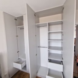 Błażej Michalak - Jasnoszara szafa wnękowa z otwartymi drzwiami, ukazująca półki i wysuwane szuflady. Widoczne okucia i drewniana podłoga. Minimalistyczny design.