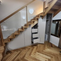 Błażej Michalak - Drewniane schody z szklaną balustradą i szarymi szafkami pod schodami z otwartymi szufladami na parkiecie w jodełkę. Widoczne oświetlenie sufitowe.