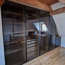 Szklana szafa wnękowa z czarnymi ramami, dopasowana do skosu poddasza. Widoczny drewniany stelaż wewnątrz i jasna drewniana podłoga.