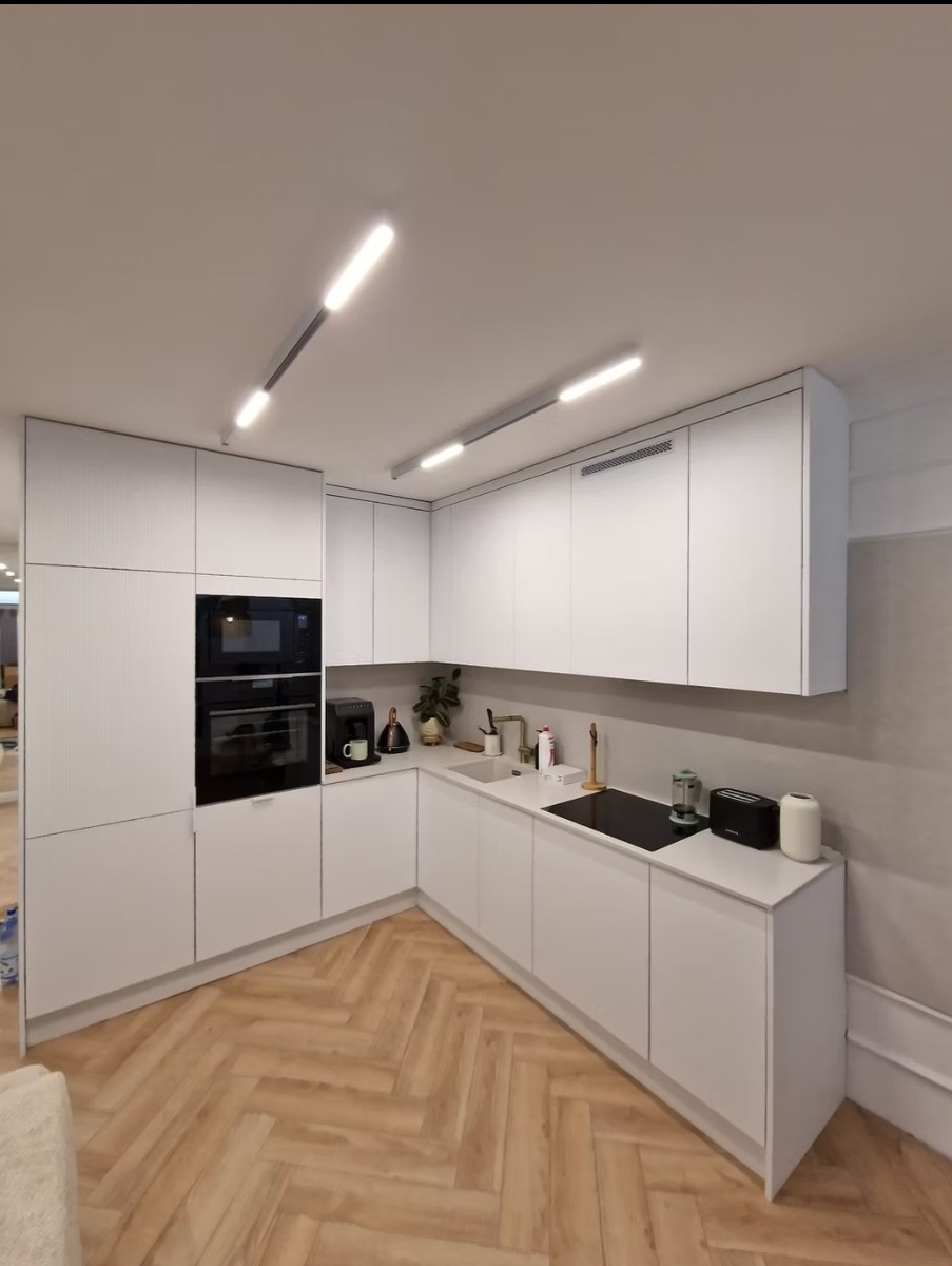 Jasna, minimalistyczna kuchnia z białymi szafkami i drewnianą podłogą w jodełkę. Oświetlenie LED podkreśla nowoczesny charakter wnętrza. Widoczny ekspres do kawy i akcesoria.
