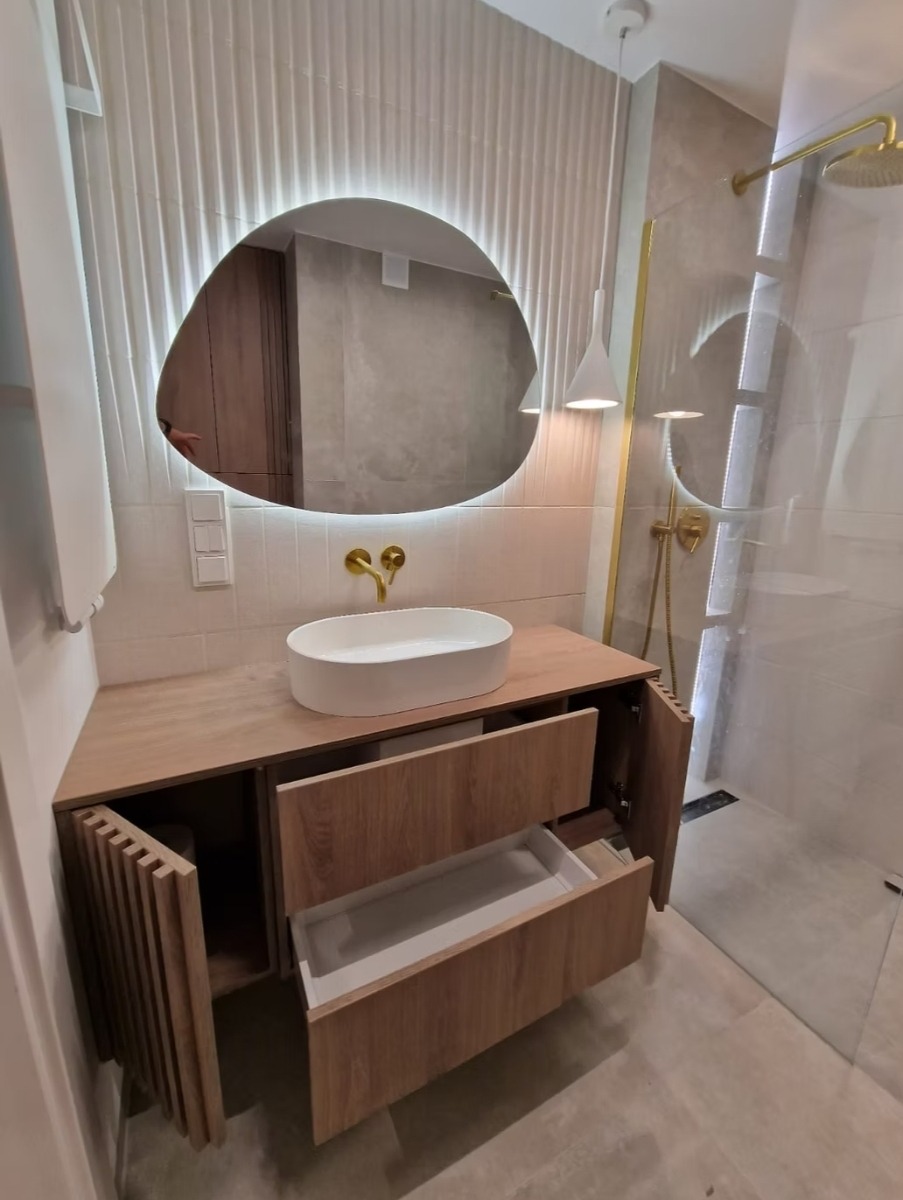 Nowoczesna łazienka z drewnianą szafką pod umywalkę, owalnym lustrem z podświetleniem LED i złotą armaturą. Widoczny otwarty front szafki i wysunięta szuflada.