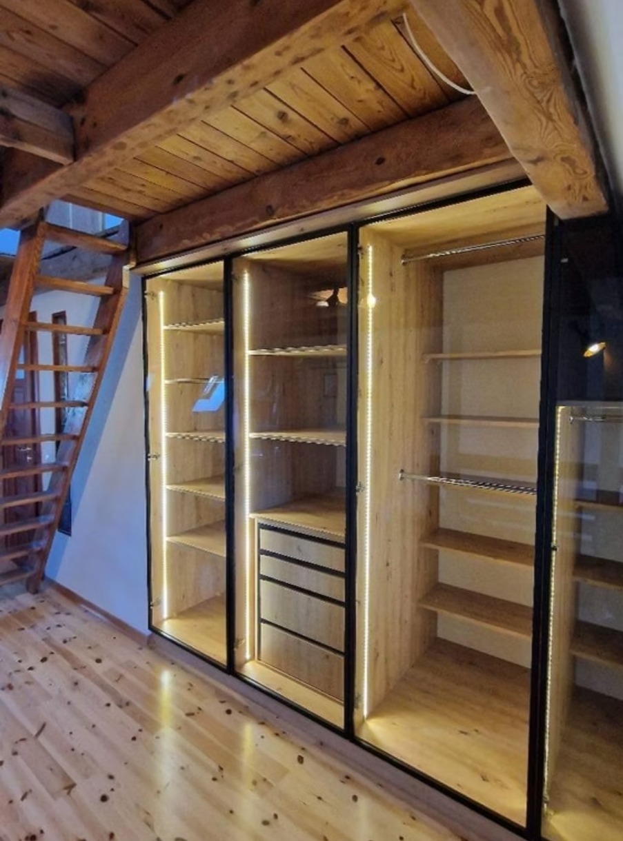Drewniana szafa wnękowa z przesuwnymi, szklanymi drzwiami, oświetlona taśmami LED, w pomieszczeniu z drewnianym stropem i schodami.