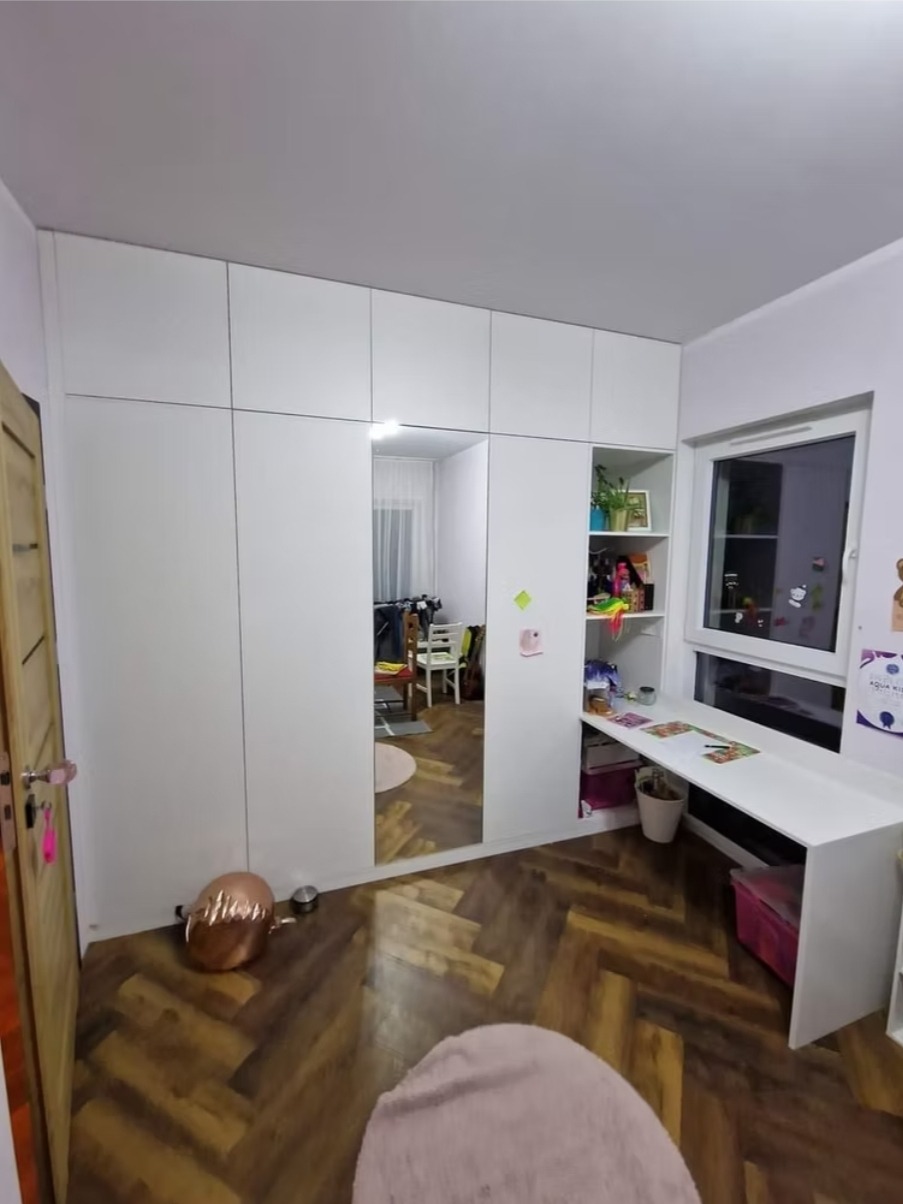 Biała szafa wnękowa z lustrem i biurkiem w pokoju dziecięcym. Podłoga w jodełkę, dekoracje na ścianach. Widok na wnętrze pokoju w lustrze.