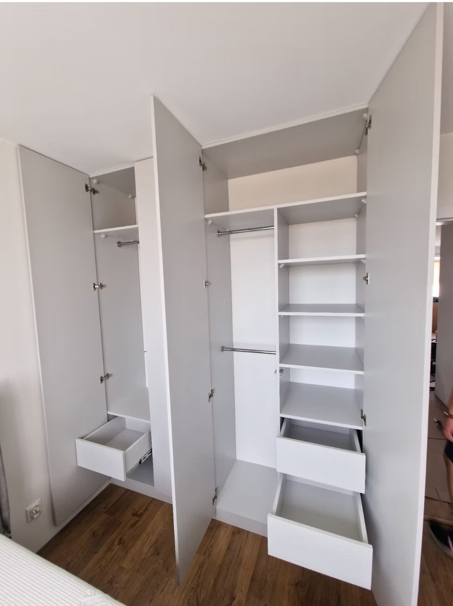 Jasnoszara szafa wnękowa z otwartymi drzwiami, ukazująca półki i wysuwane szuflady. Widoczne okucia i drewniana podłoga. Minimalistyczny design.