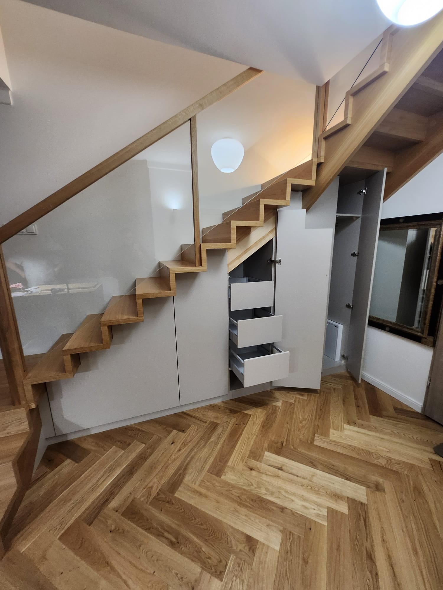 Drewniane schody z szklaną balustradą i szarymi szafkami pod schodami z otwartymi szufladami na parkiecie w jodełkę. Widoczne oświetlenie sufitowe.