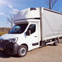 Biały bus Renault z plandeką na tle błękitnego nieba i drzew bez liści. Pojazd zaparkowany na asfaltowej drodze, widoczny z boku pod lekkim kątem.