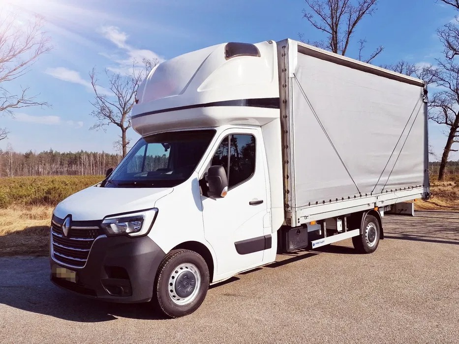 Biały bus Renault z plandeką na tle błękitnego nieba i drzew bez liści. Pojazd zaparkowany na asfaltowej drodze, widoczny z boku pod lekkim kątem.