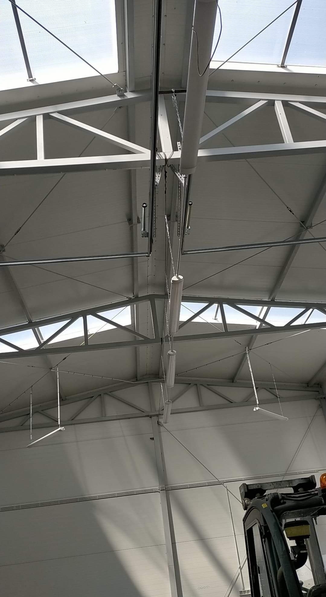 Wnętrze hali z widoczną konstrukcją dachu, oświetleniem przemysłowym i elementami instalacji elektrycznej oraz fragmentem wózka widłowego w prawym dolnym rogu.