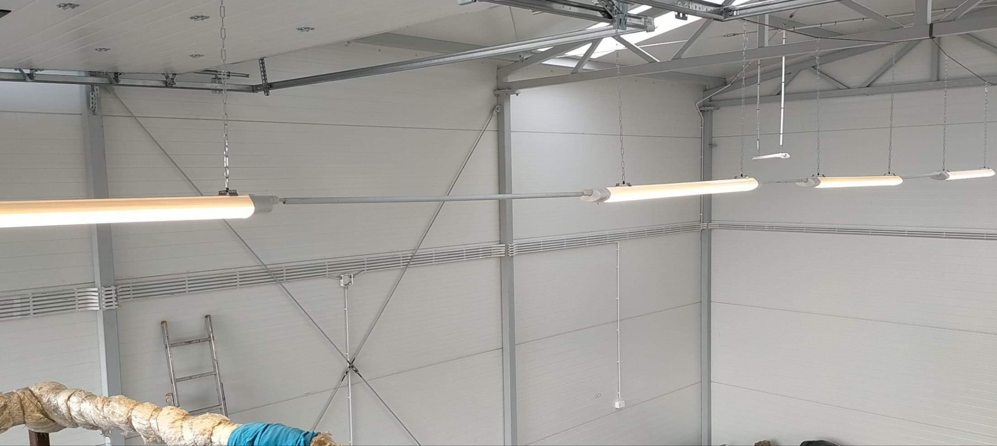 Oświetlenie LED w hali: rzędy lamp rurowych zawieszonych na łańcuchach, równomiernie rozmieszczone, oświetlające przestrzeń magazynową z metalową konstrukcją.