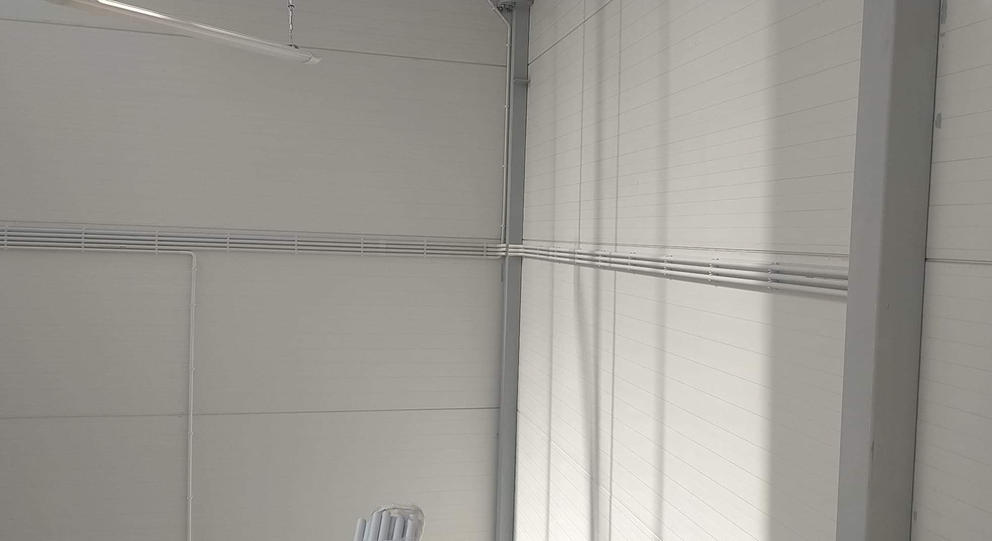 Białe rury elektryczne ułożone poziomo wzdłuż białej ściany z panelami w hali przemysłowej, widoczne elementy konstrukcyjne budynku.