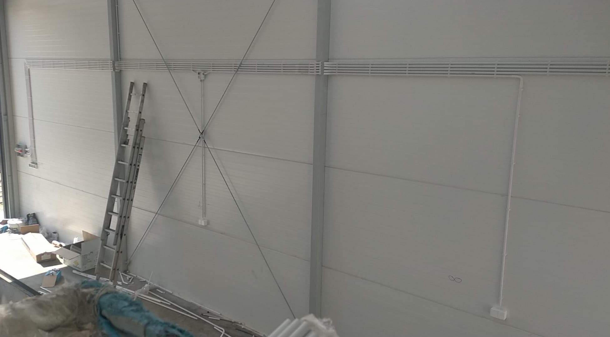 Wnętrze hali z widoczną instalacją elektryczną w białych rurkach, drabina oparta o ścianę. Widok z góry na zamontowane elementy.