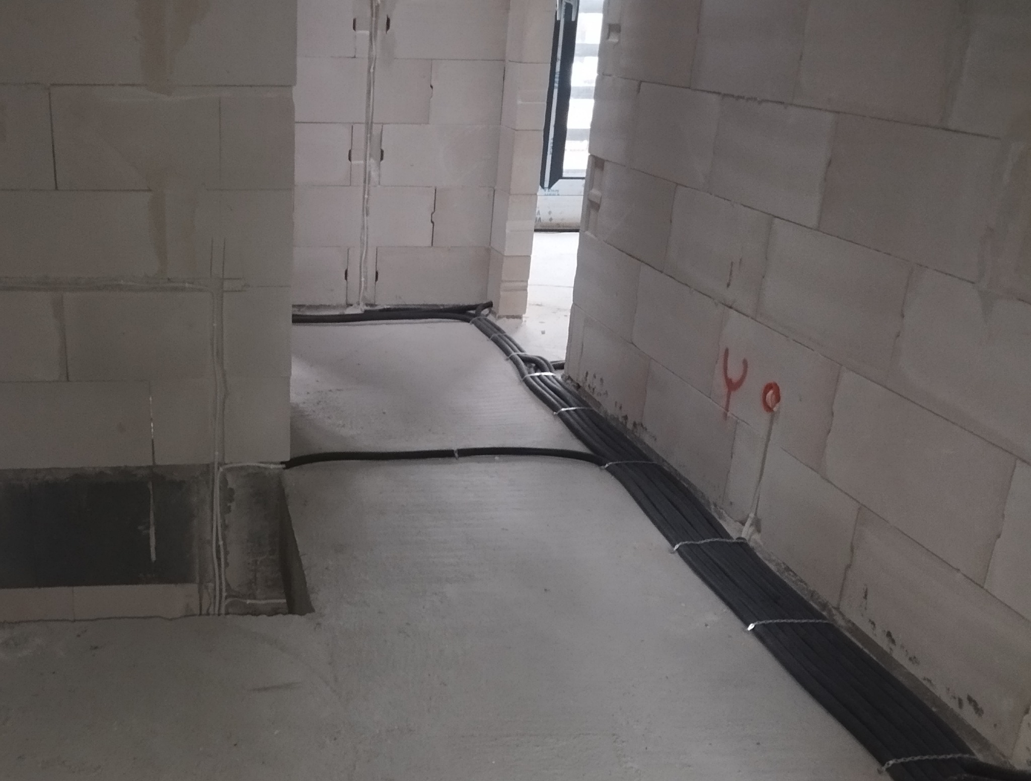 Wiązka czarnych kabli prowadzona po szarej posadzce w surowym wnętrzu budynku z białych bloczków, widoczne oznaczenia na ścianie, wstępny etap instalacji.