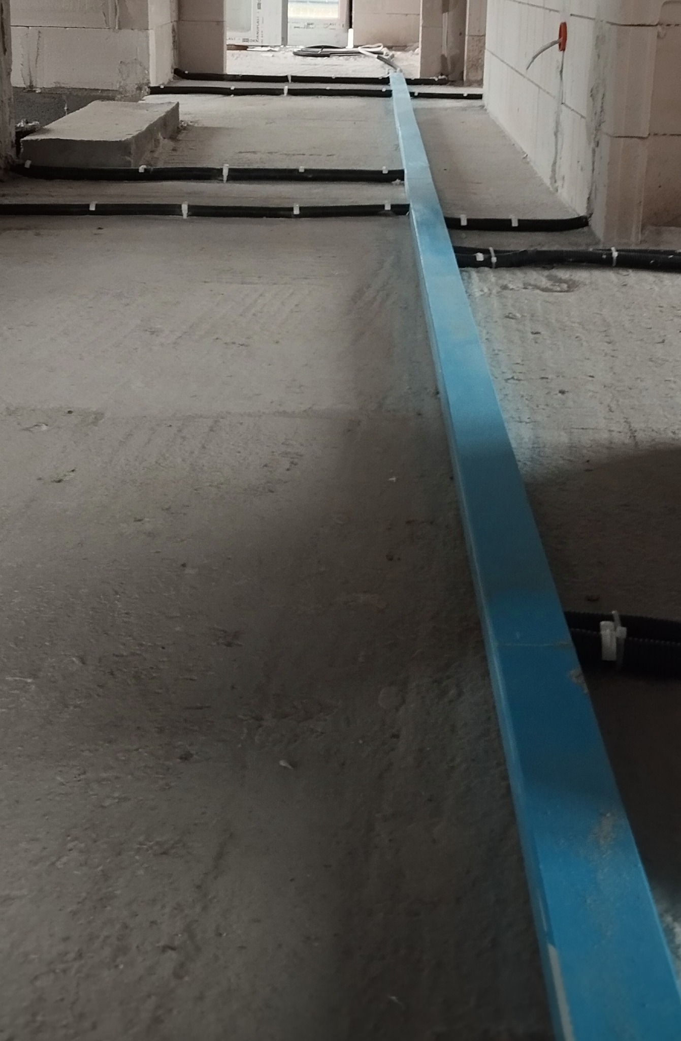 Podłogowy montaż rur w surowym wnętrzu. Czarna rura przymocowana białymi uchwytami do podłoża. Niebieska listwa wzdłuż ściany. Etap instalacji w budynku.