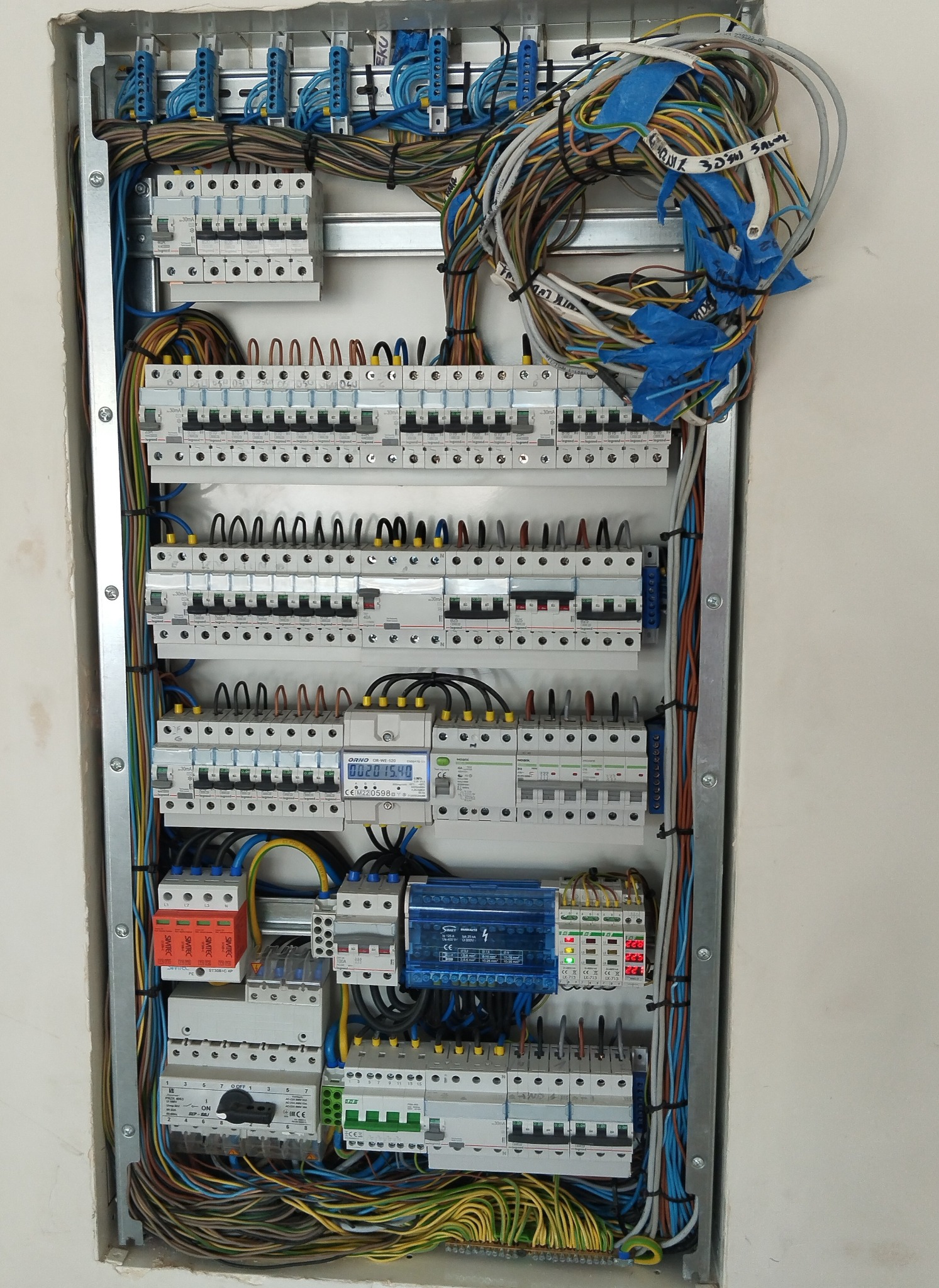 Rozdzielnica elektryczna w metalowej obudowie, z licznymi zabezpieczeniami, przewodami i elementami pomiarowymi. Widoczne kable oznaczone taśmą i opisane.