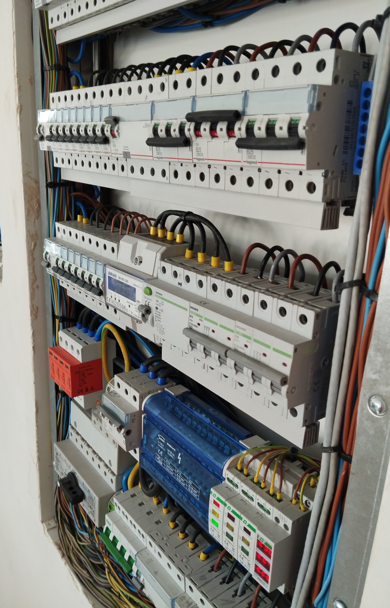 Szczegółowe ujęcie rozdzielnicy elektrycznej z bezpiecznikami, przewodami i elementami zabezpieczającymi, zamontowanej wewnątrz budynku, z widocznymi oznaczeniami producentów.