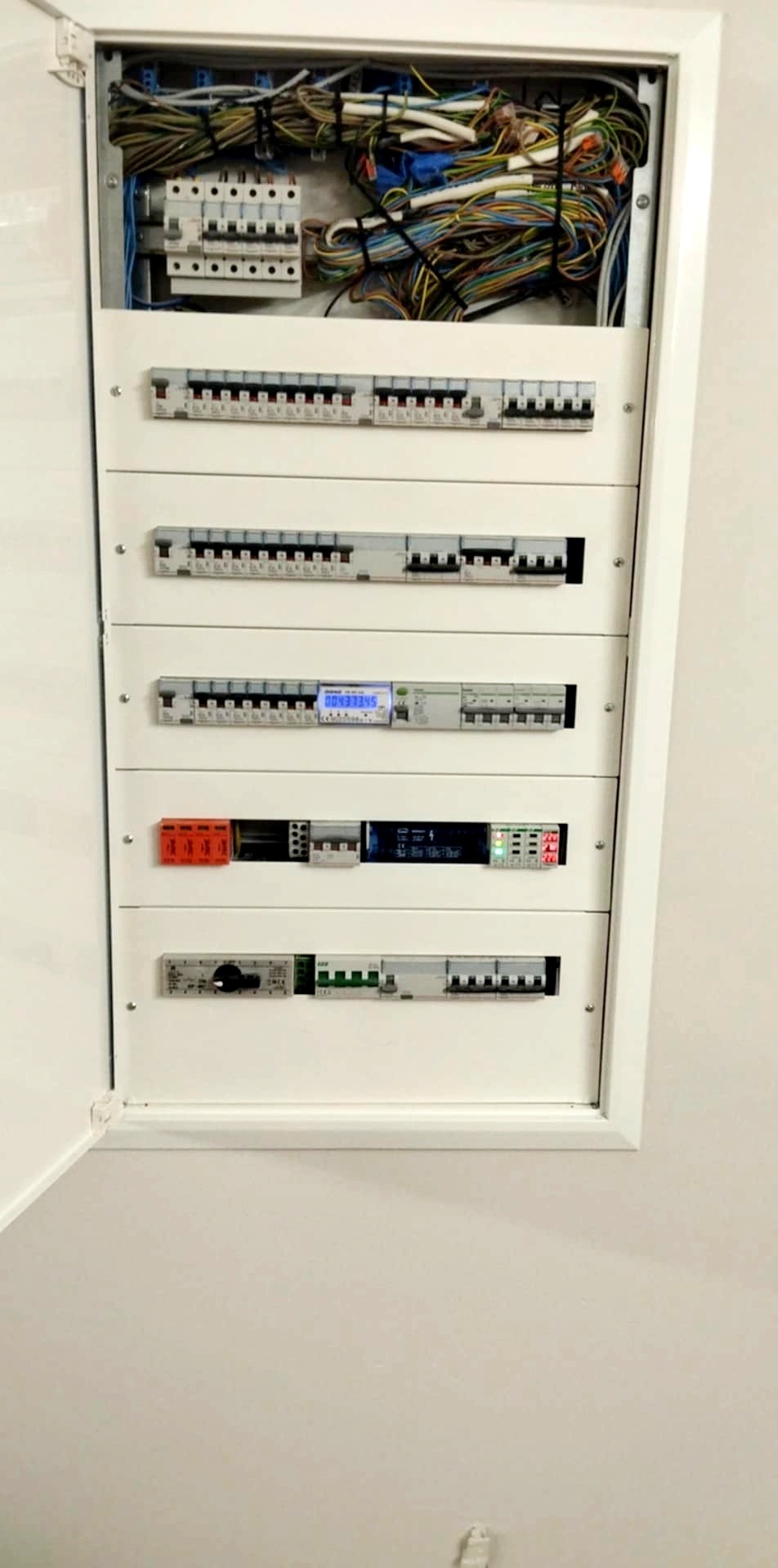 Rozdzielnica elektryczna w białej obudowie, widoczne bezpieczniki, kable i liczniki. Zastosowano różne typy zabezpieczeń.