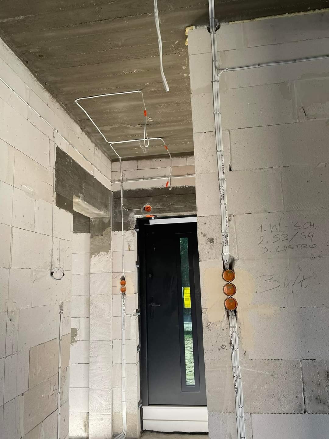 Surowy stan: białe kable elektryczne prowadzone po ścianach z betonu komórkowego, widoczne puszki instalacyjne i czarne drzwi wejściowe w nowym budynku.