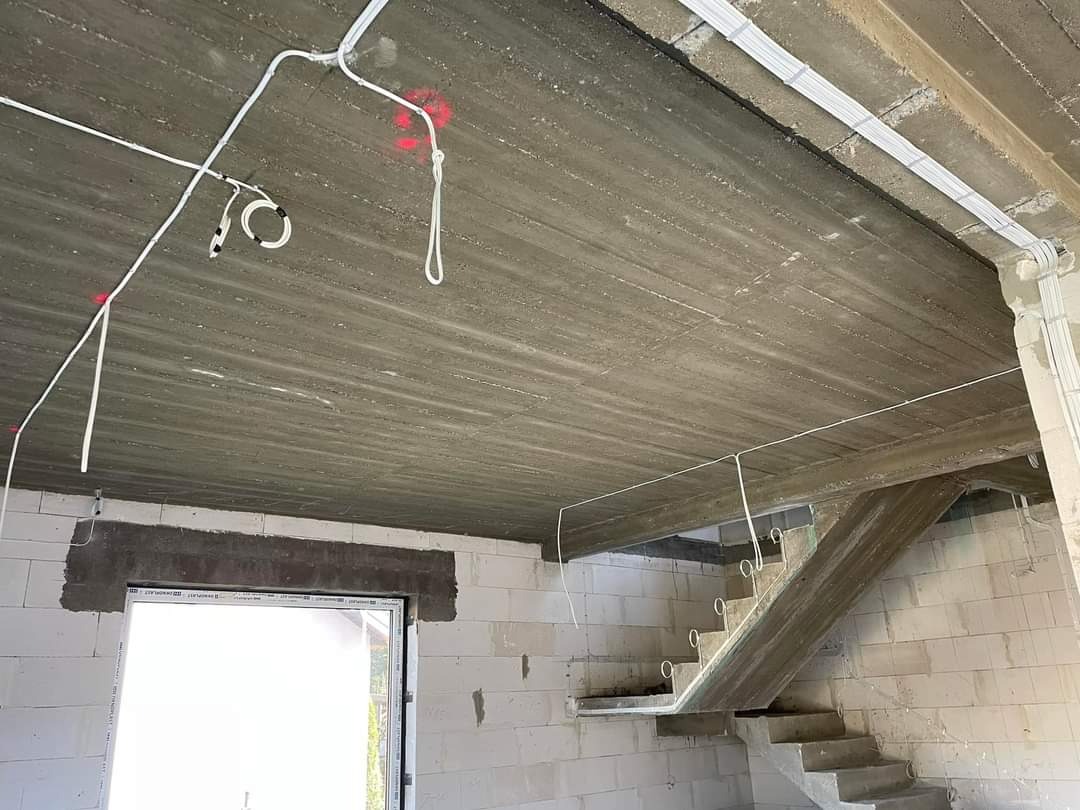 Surowy strop betonowy z instalacją elektryczną w białych rurkach, czerwone znaczniki, ściany z białych bloczków, schody betonowe w stanie surowym.
