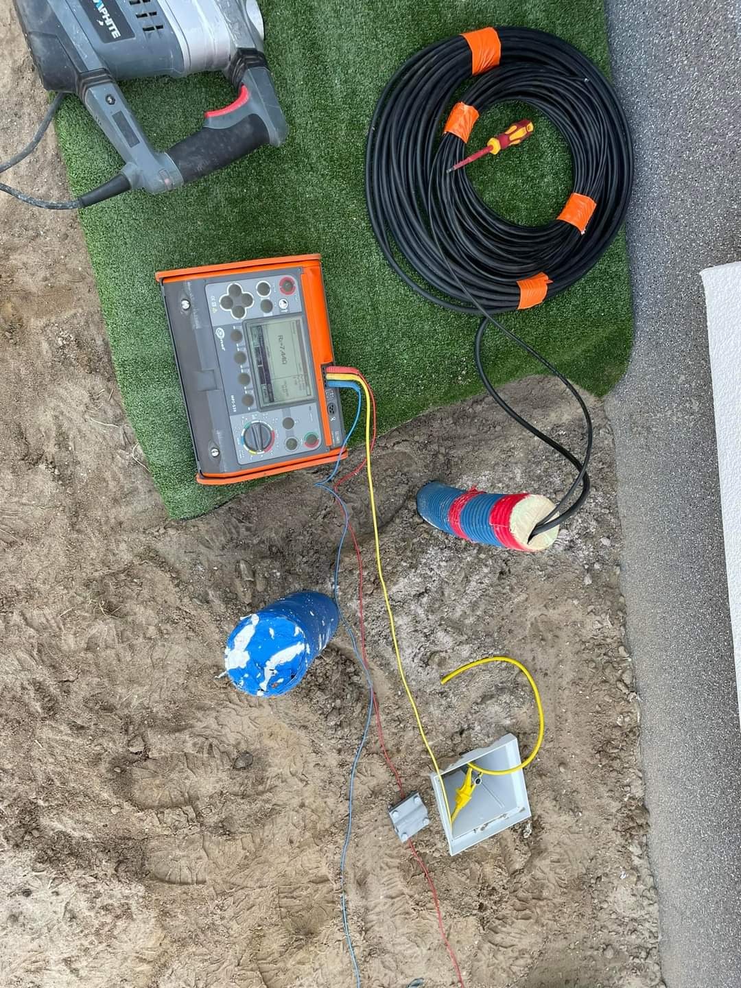 Miernik, przewody i wiertarka na trawie i ziemi. Przygotowanie do pomiarów elektrycznych, widoczne elementy instalacji w ziemi.